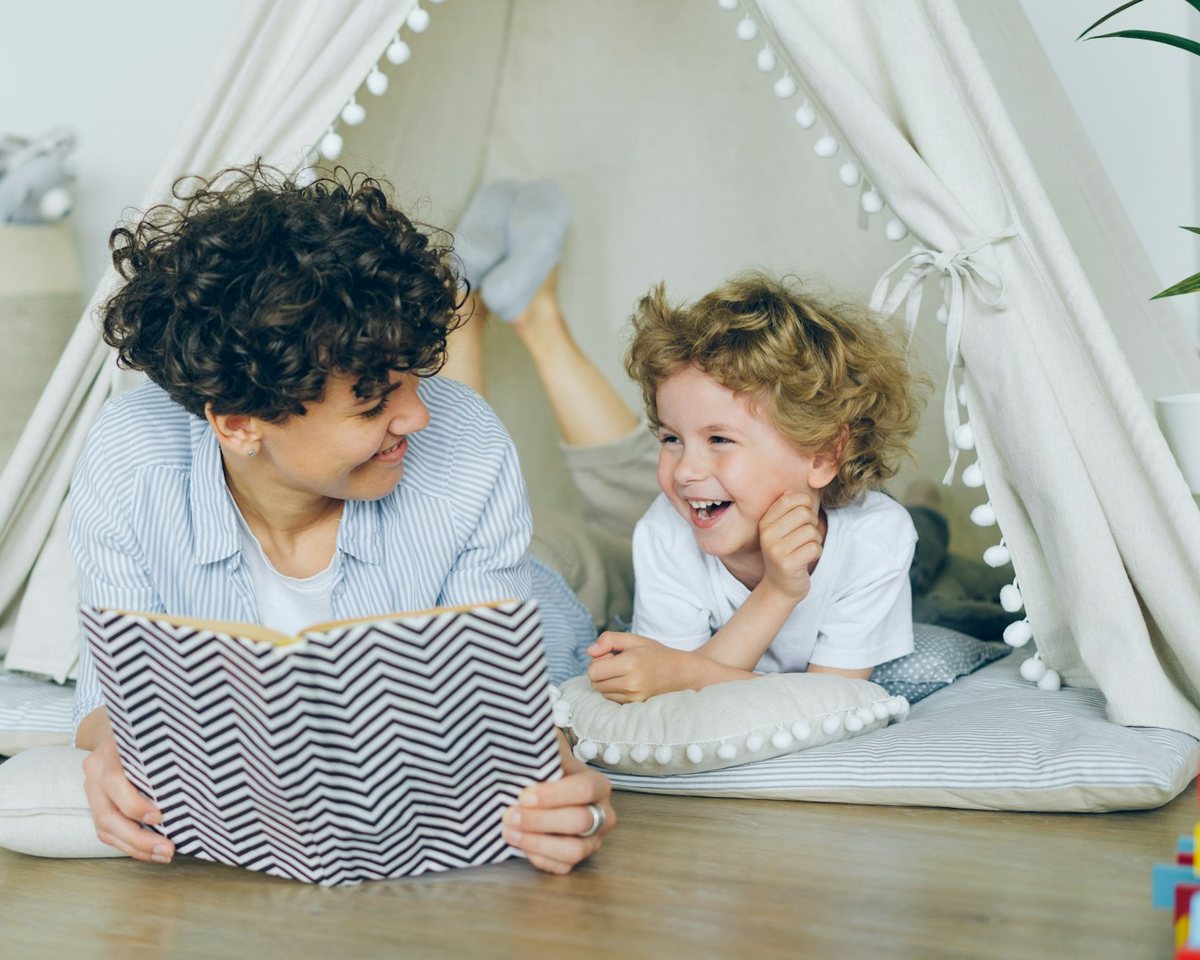 A mother reading a book to her laughing son inside a blanket tent