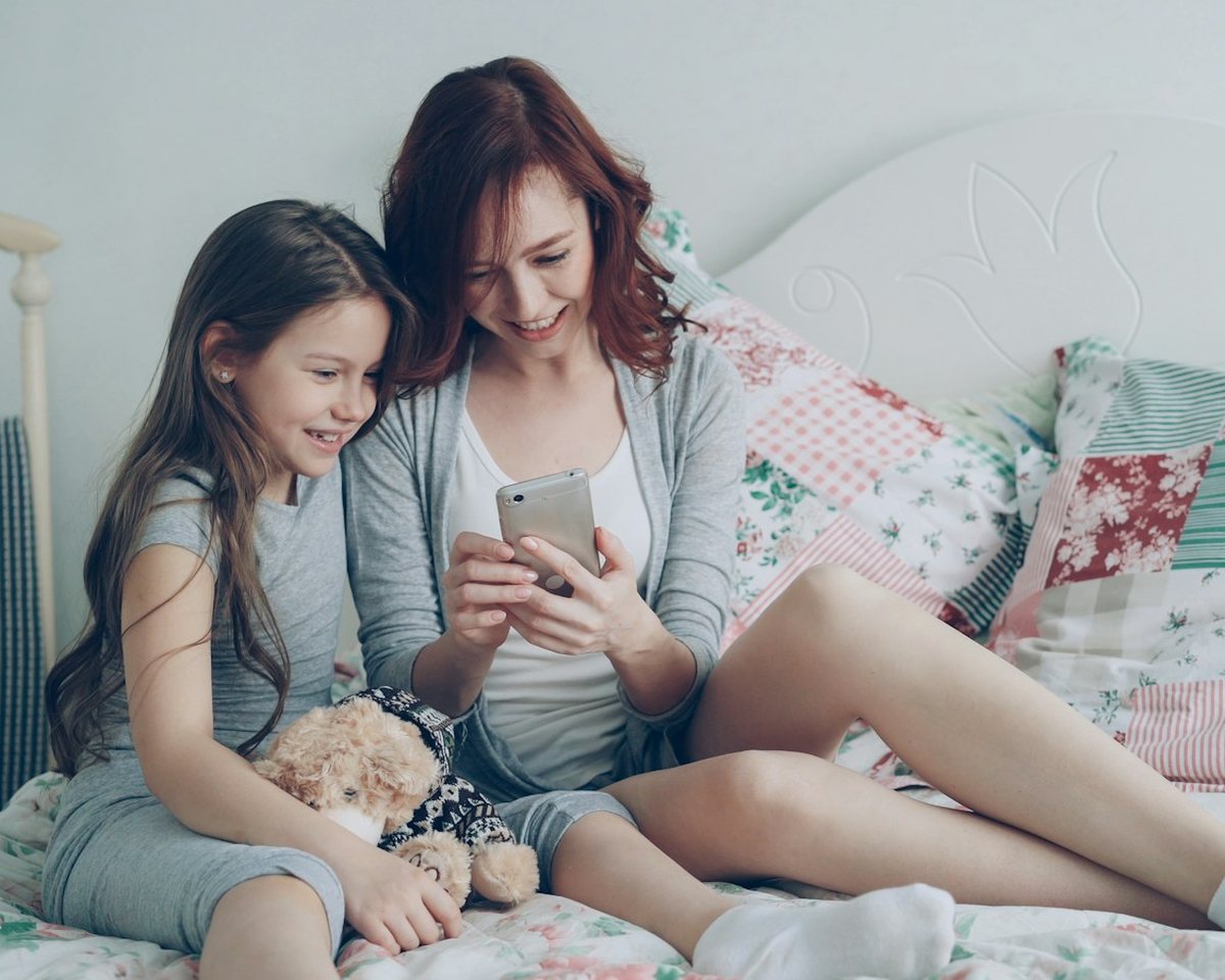 A mother and her young daughter sitting on a bed and smiling at a phone together